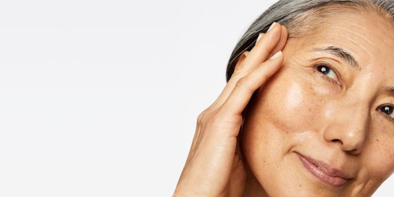A close-up photograph of a smiling woman with grey hair gently touching her temple, showcasing a radiant, dewy, and glowing complexion with natural skin texture against a clean, off-white background.