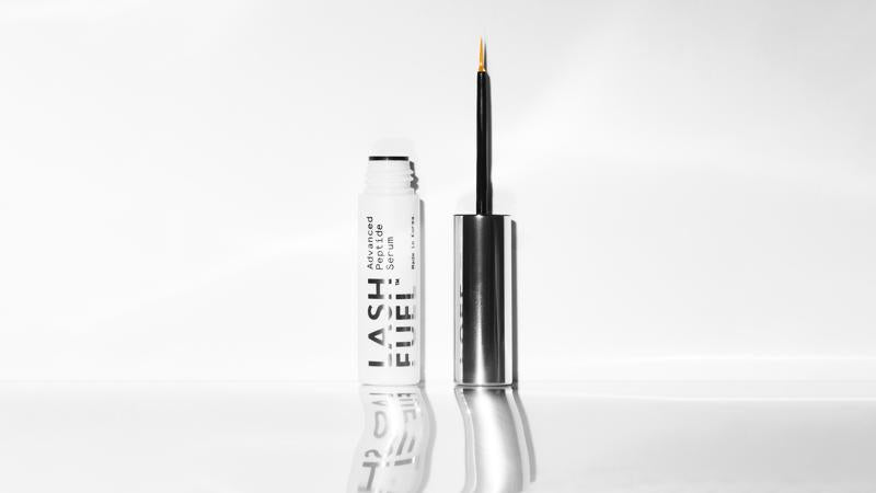 Beauty Pie Lash Fuel Advanced Peptide Serum white bottle and metallic silver cap standing upright, showing a fine-tipped applicator brush. The product reflects on a glossy white surface against a clean, minimal white background.