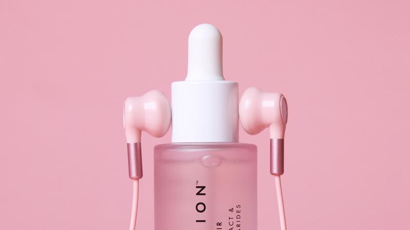 A close-up of a Beauty Pie Japanfusion serum dropper bottle styled with a pair of pink earbuds on either side of its white cap, set against a solid pastel pink background.