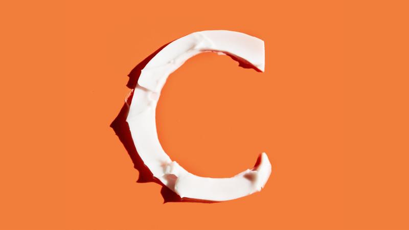 A thick swatch of white skincare cream shaped like a capital letter "C" centered on a solid, bright orange background.